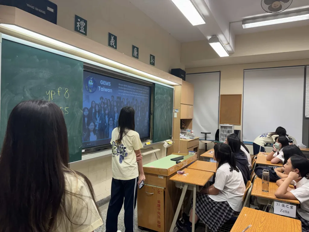 A team member at the front of the classroom beside a projected GEMS Taiwan team photograph.