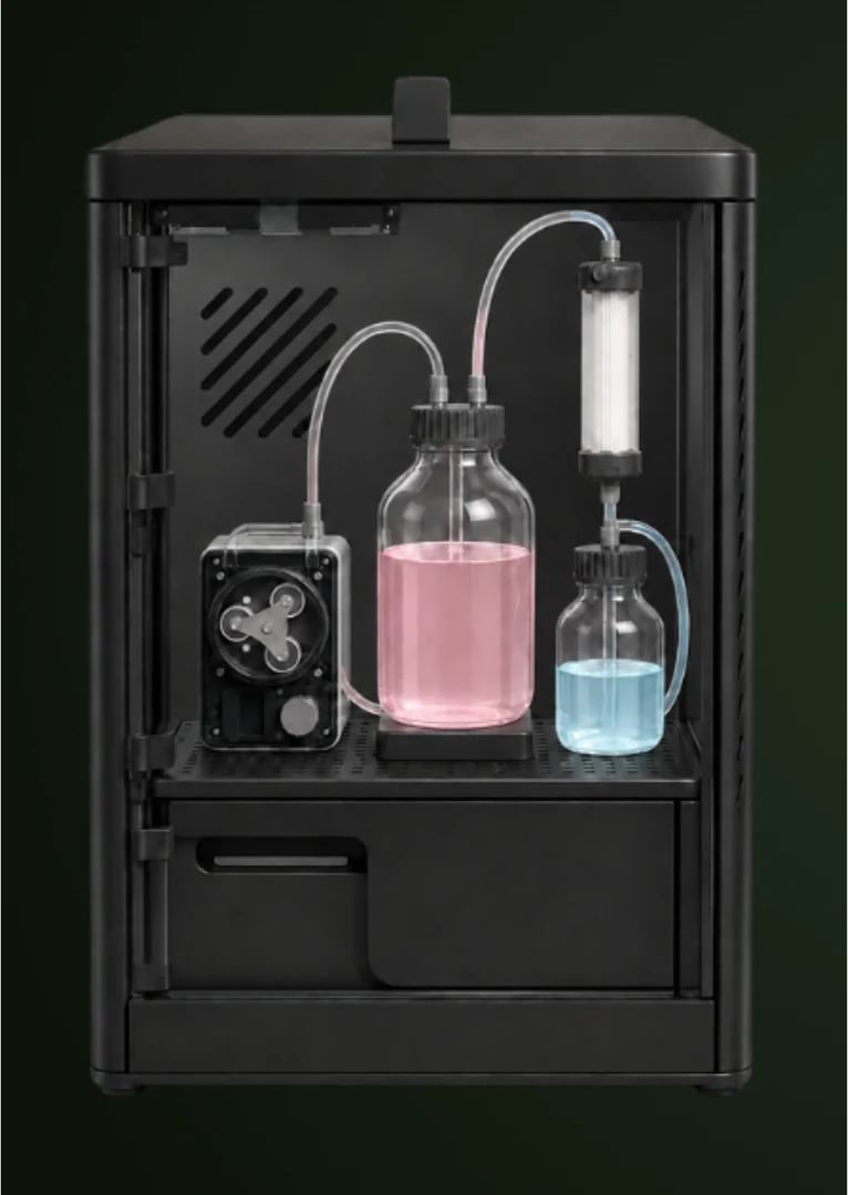 A render of the bioreactor product: a black cabinet with an open front holding a pump, a bottle of pink culture, a smaller bottle of blue liquid and a hollow fiber cartridge joined by tubing.