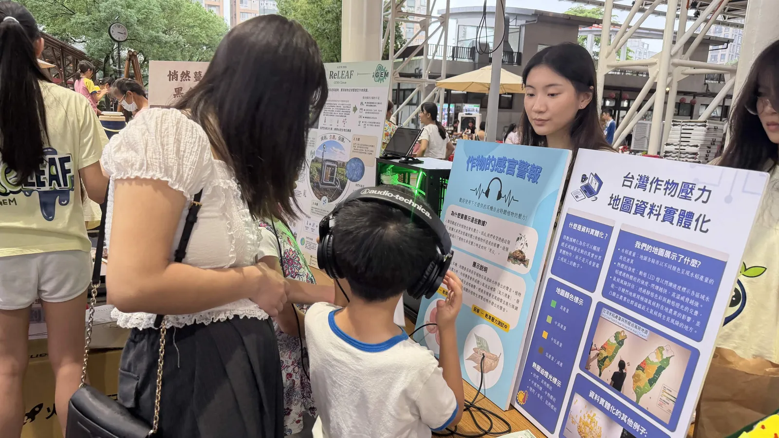 A child wearing headphones listens beside a poster about Taiwan&rsquo;s crop stress map.
