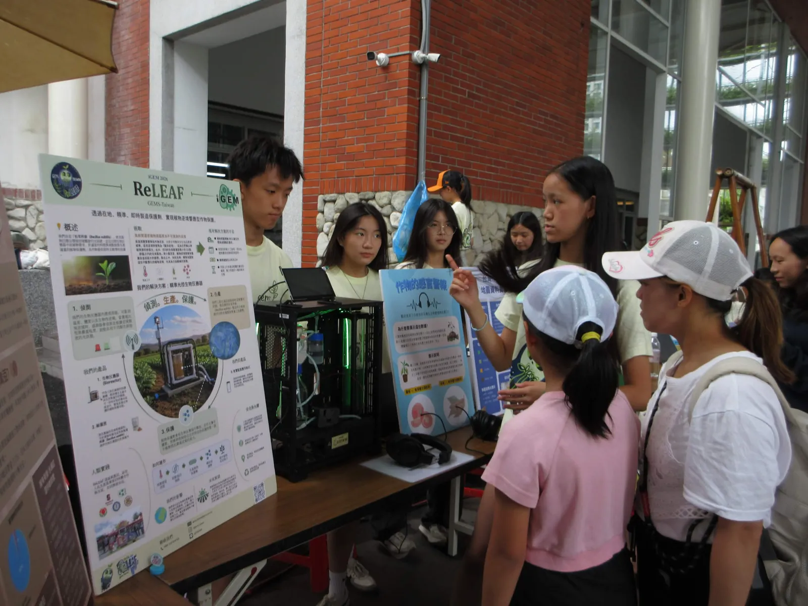 Visitors in front of a ReLEAF poster board with the bioreactor prototype lit green on the table.