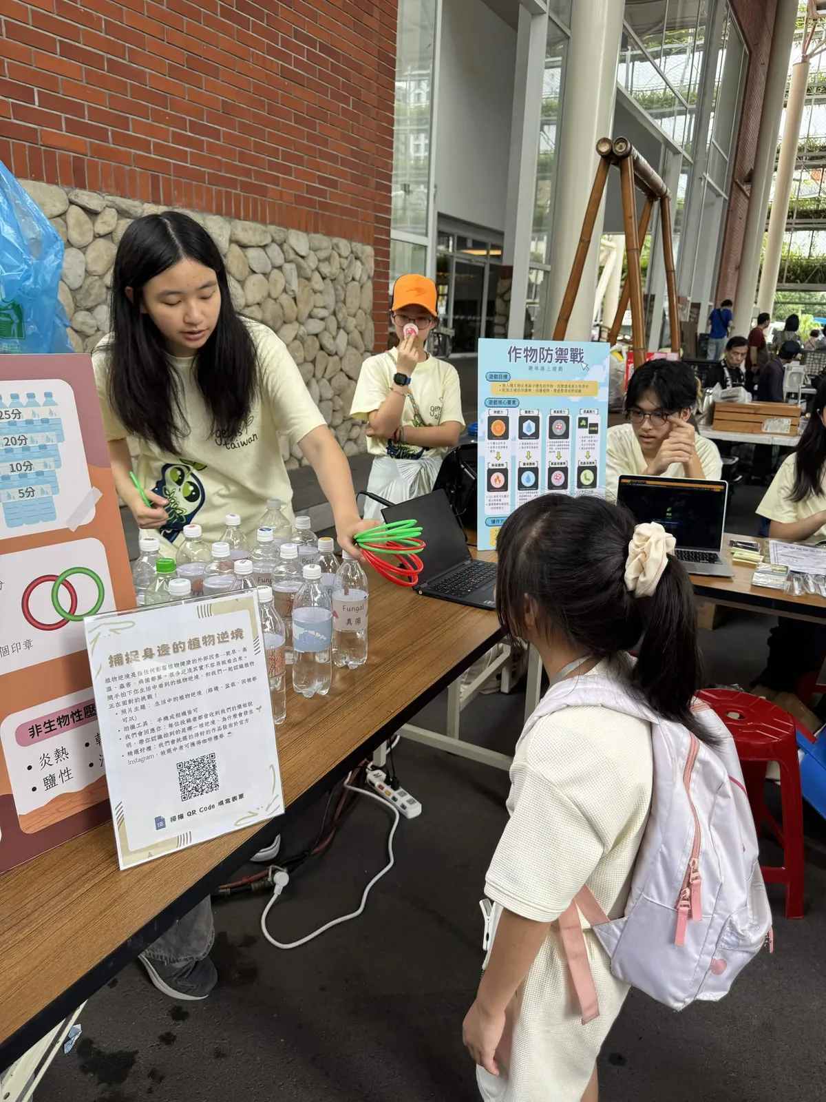 A girl aims a ring at bottles on a table while a student watches.