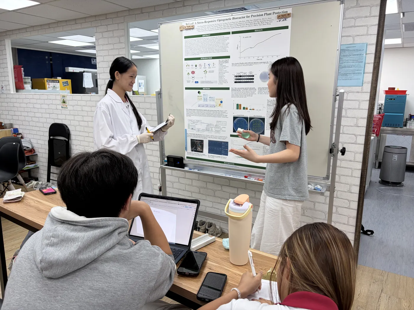 Two students rehearsing in front of a printed poster in the lab while teammates watch.