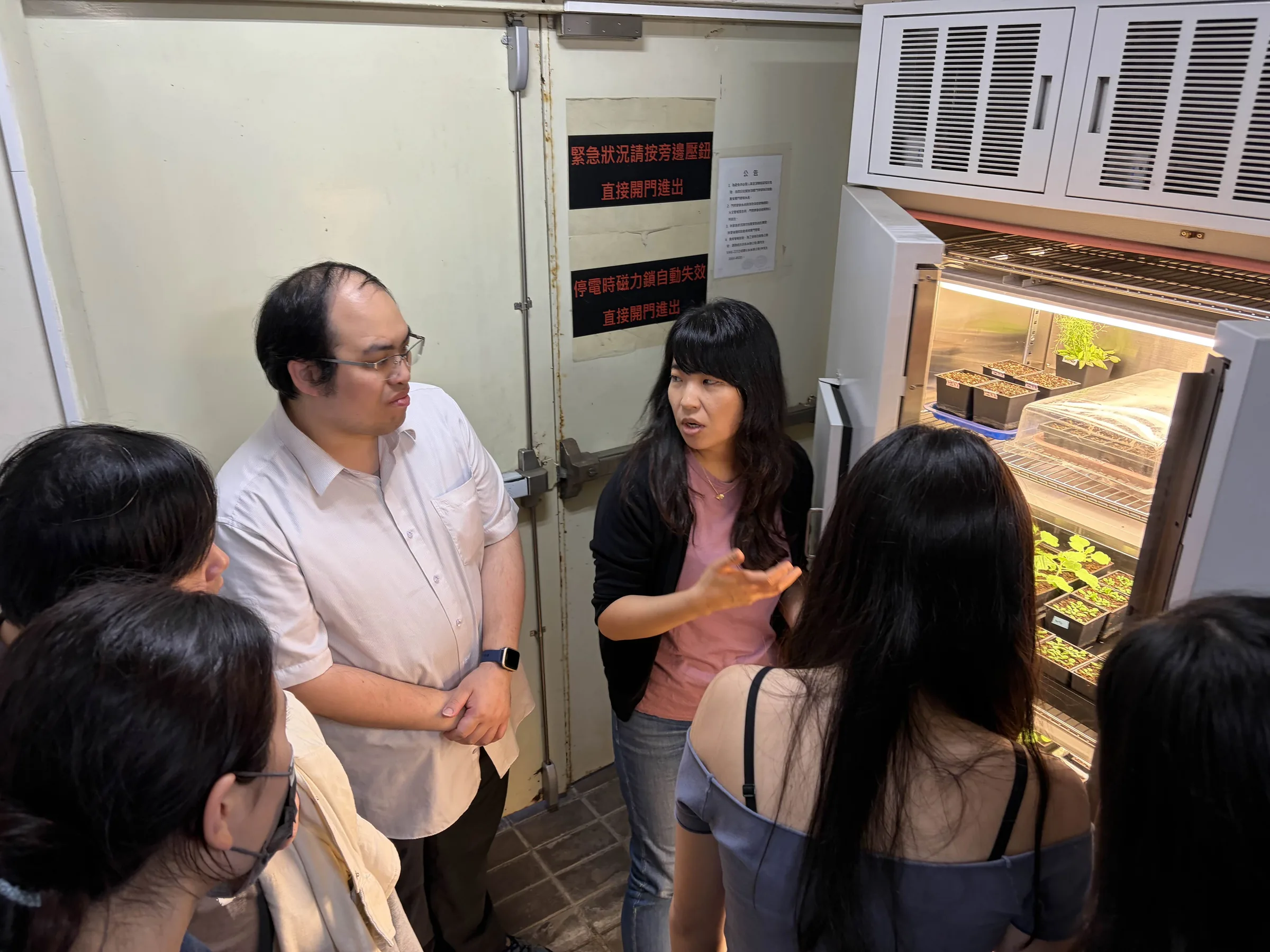 Prof. Cheng talking with a teacher and students beside an open growth chamber holding trays of seedlings.