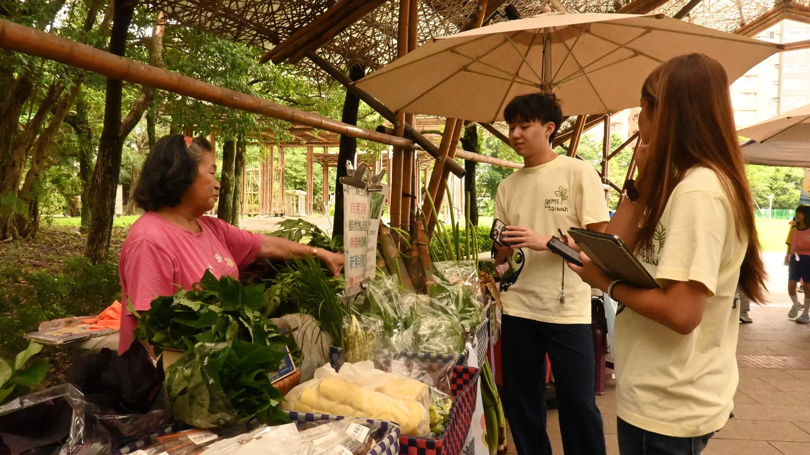 A student with a clipboard interviewing a woman at a vegetable stall under trees.