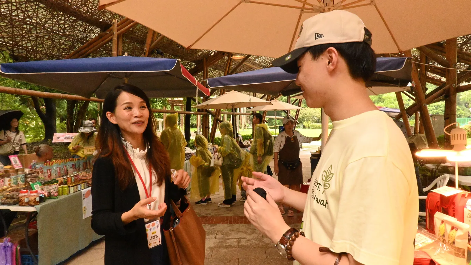 A student in a cap talking with a woman wearing a lanyard at the market.
