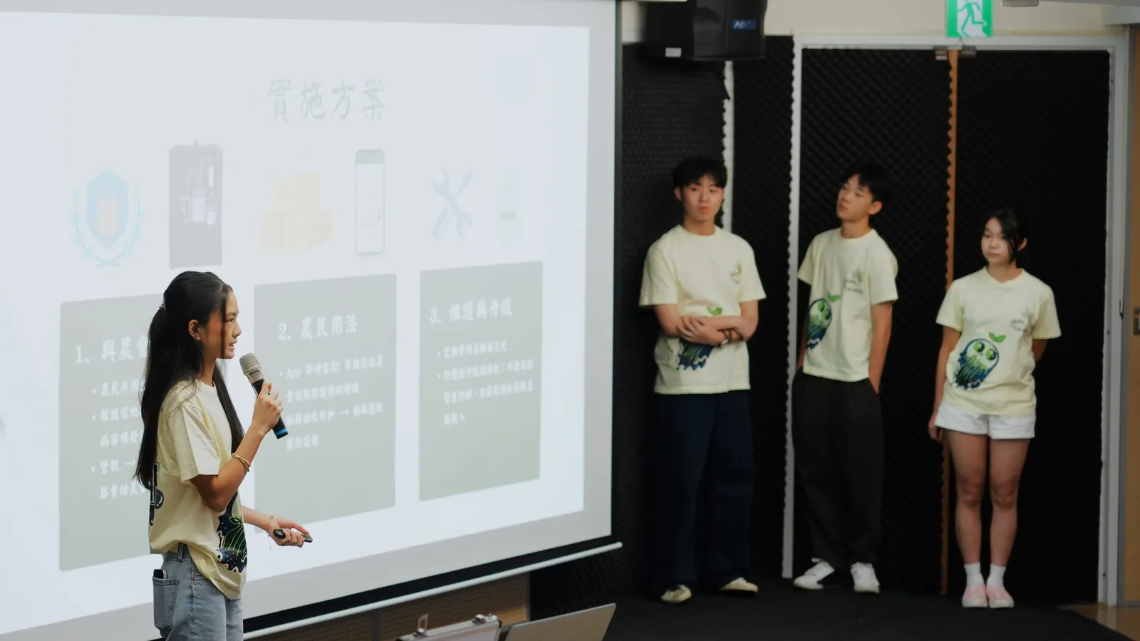 Four students presenting on stage beside a projected slide.