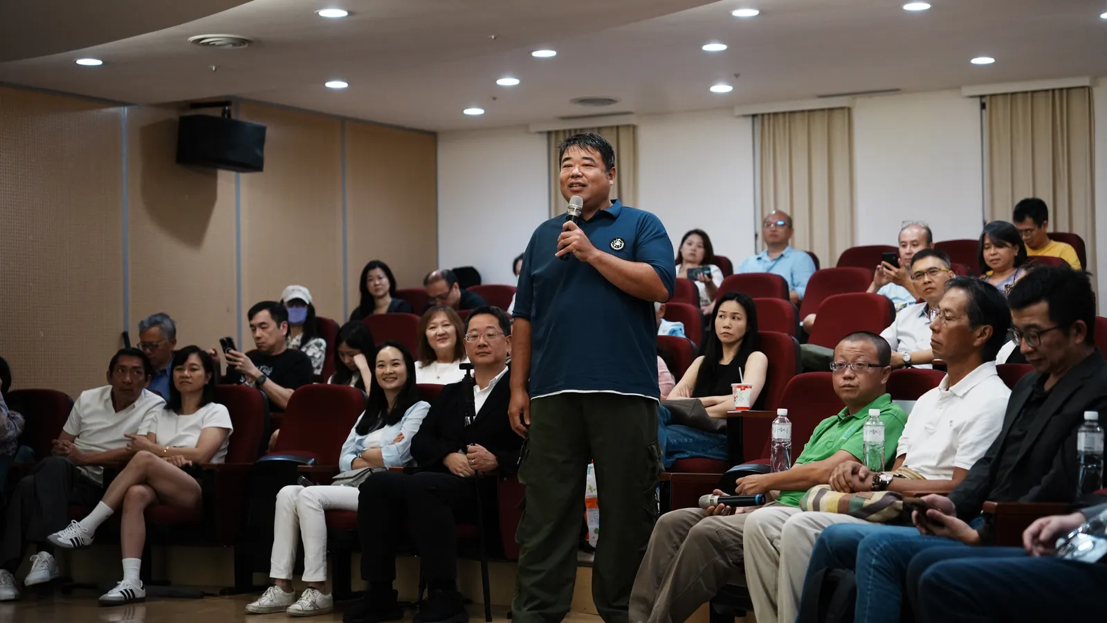 A farmer standing in the audience holding a microphone while others listen.
