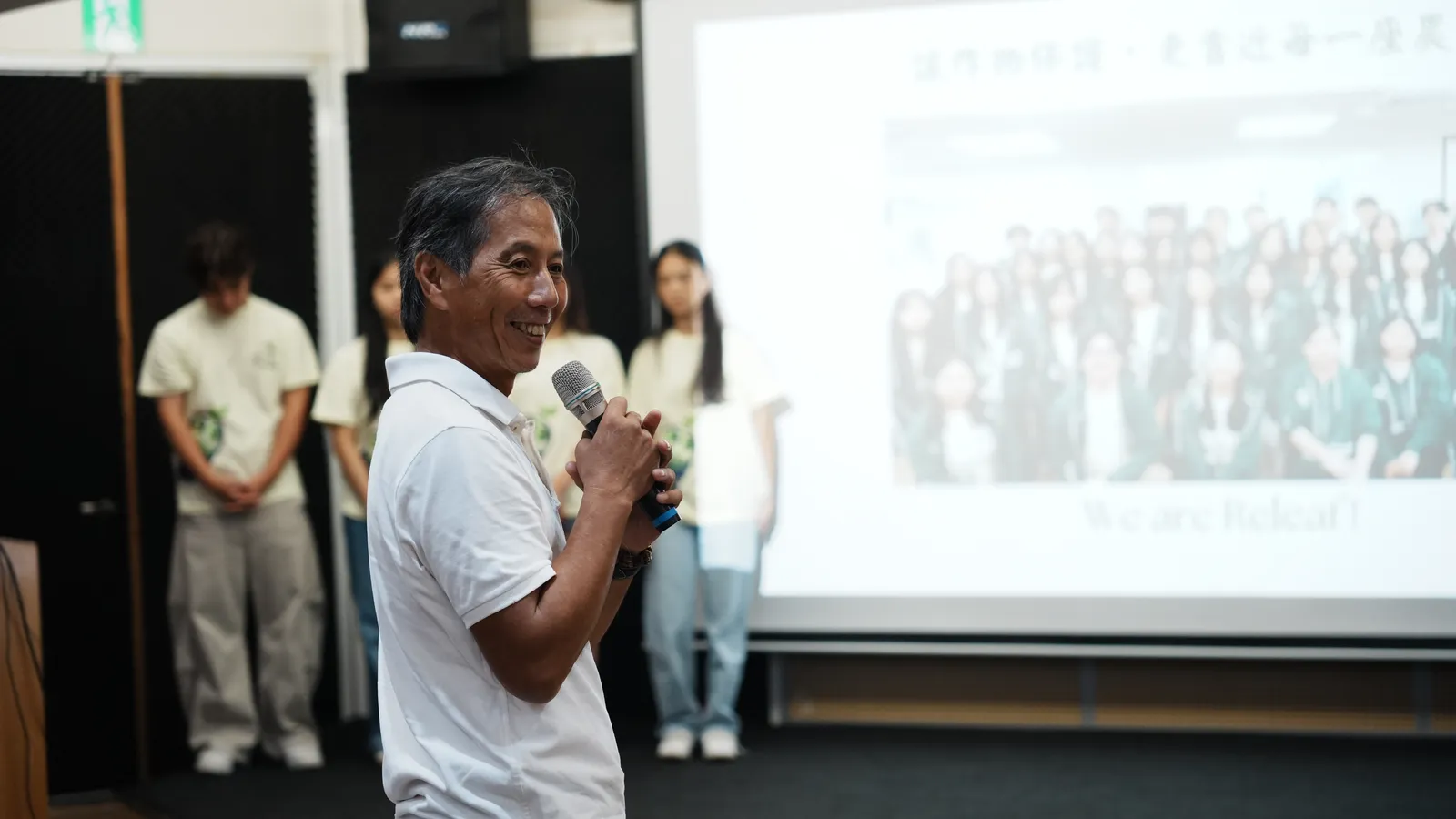 An older farmer in a white shirt speaking into a microphone near the stage.